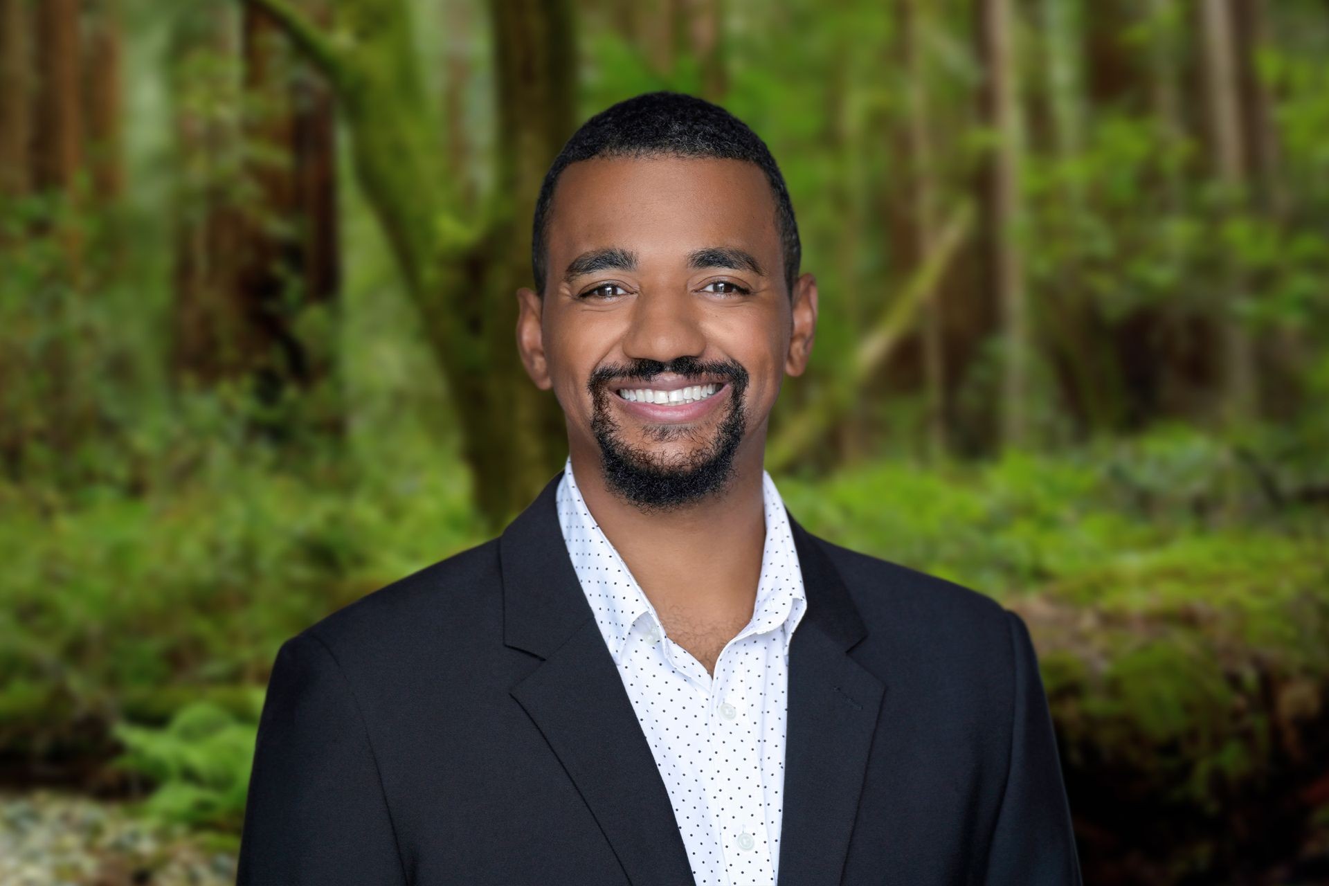 Man in a black blazer and white shirt smiling in front of a lush green forest background.