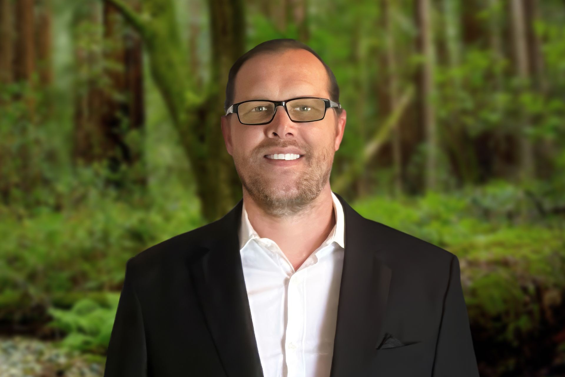 Man in a black suit and white shirt standing in front of a lush, green forest background.