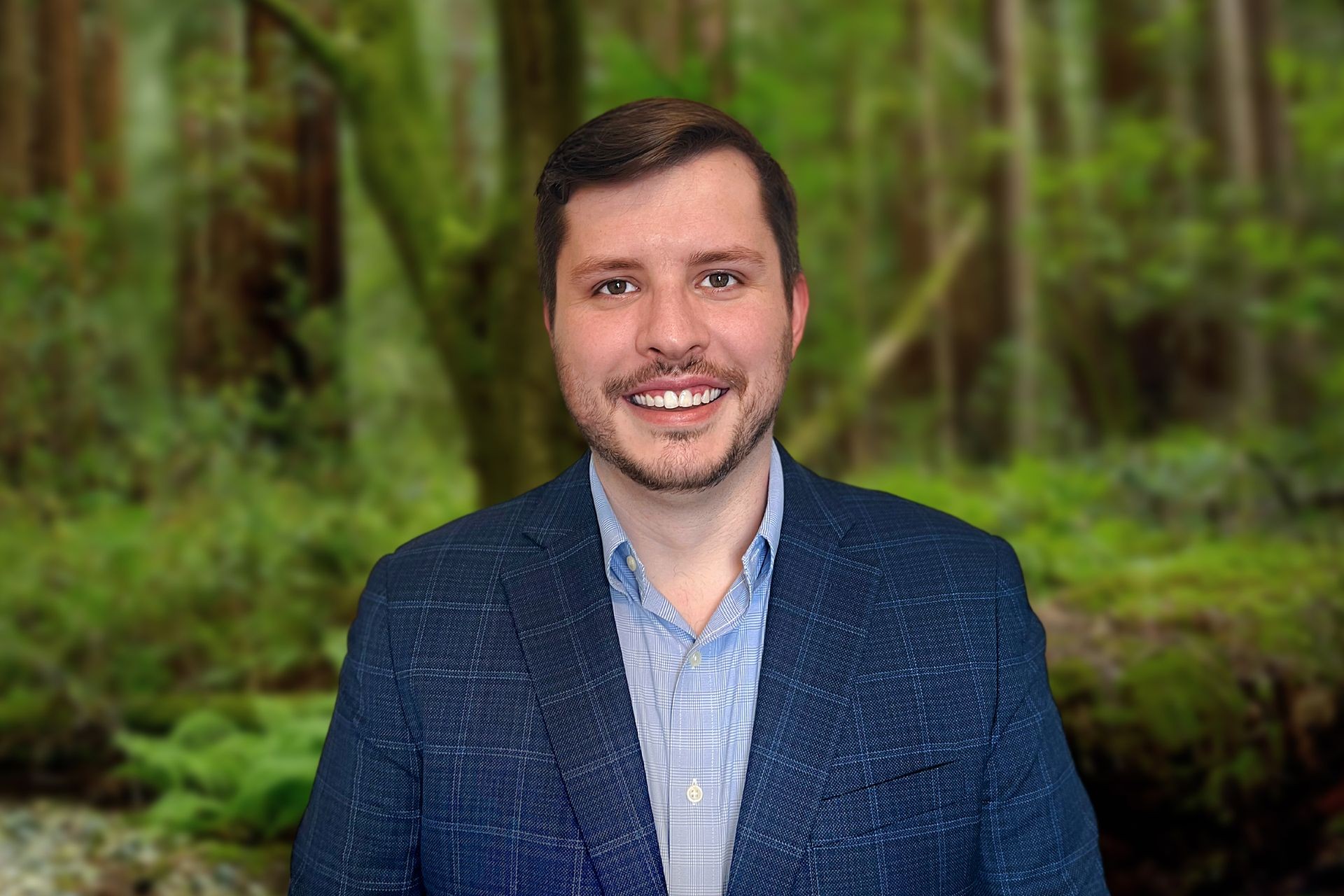 Man wearing a blue blazer and light blue shirt, smiling in front of a blurred forest background.