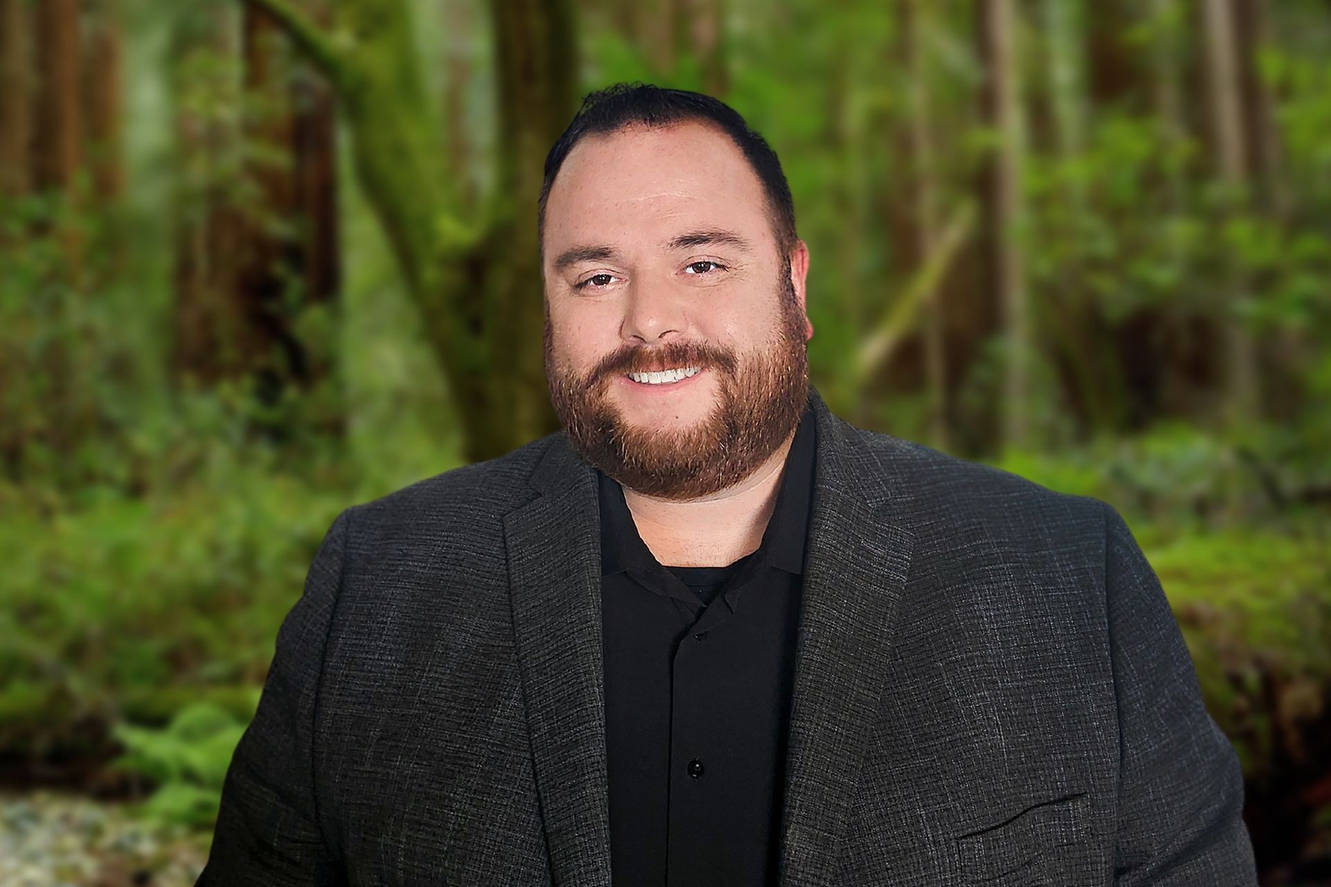 Man in a dark suit jacket and black shirt standing outdoors with a blurred green forest background.